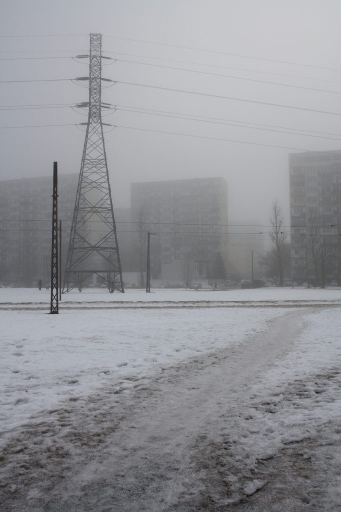 Widok słupa wysokiego napięcia, w tle mieszkalne wieżowce na osiedlu Retknia w Łodzi, całość krajobrazu skrywa się w mgle